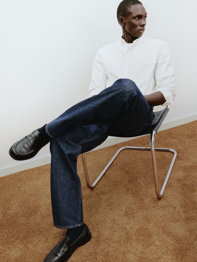 Black man seated in a chrome chair wearing a white shirt, dark blue jeans, and black loafers against a white wall and brown carpet.