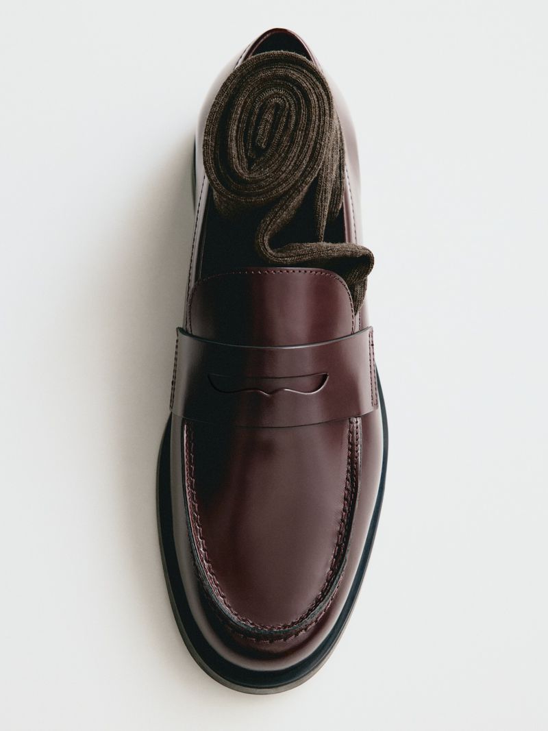 Top-down view of a glossy burgundy leather penny loafer with a rolled brown knit sock tucked inside on a white background.