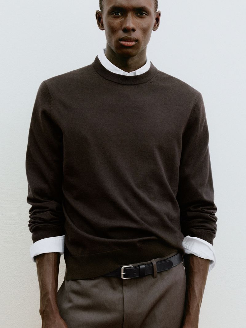 Black male model wearing a dark brown crewneck sweater layered over a white collared shirt against an off-white backdrop.