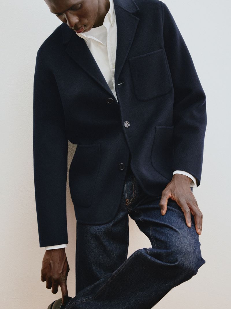 Black male model wearing a dark navy wool blazer, white collared shirt, and dark denim jeans against a white backdrop.
