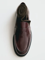 Top-down view of a glossy burgundy leather penny loafer with a rolled brown knit sock inside on a clean white background.