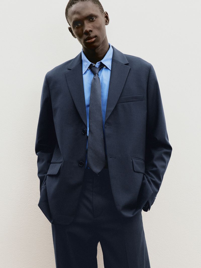 Black male model in a tailored navy blue suit, blue collared shirt, and dark tie standing against a plain white background.