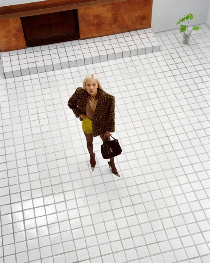 High-angle shot of Rosé wearing a tweed blazer and holding a black handbag on a white tiled floor.