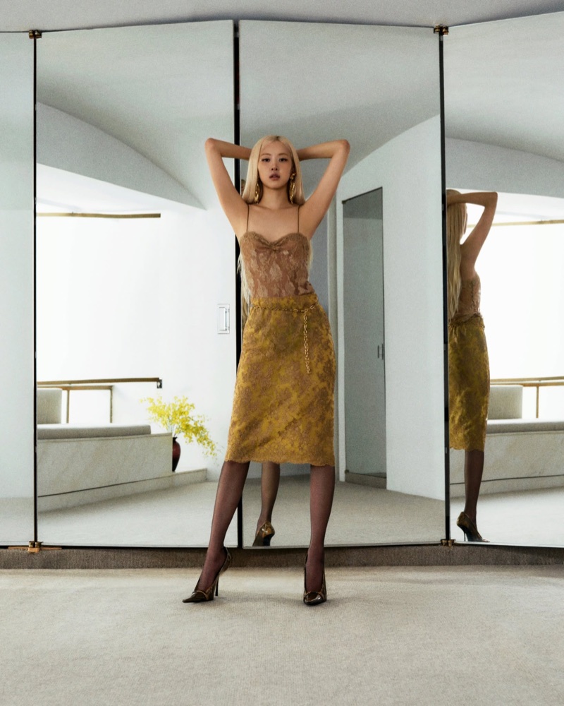 ROSÉ in a patterned gold dress and sheer black stockings standing in front of three panel mirrors for Yves Saint Laurent.