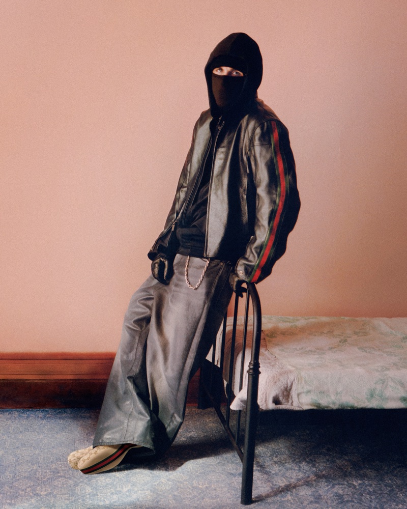 Model wearing a black balaclava, hooded leather jacket, and wide trousers leaning against a metal bed frame.