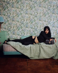 Woman in black outfit reclining on a green bed against floral wallpaper with a black handbag for Gucci Primavera 2026