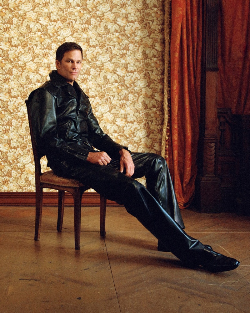 Tom Brady seated on a wooden chair wearing a black leather jacket and matching leather trousers in a wallpapered room.