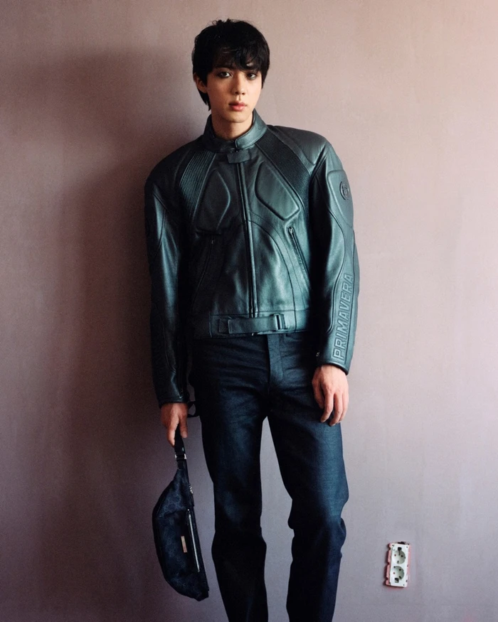Jin posing in a dark leather motorcycle jacket and dark trousers against a wall, holding a small bag for Gucci Primavera 2026.