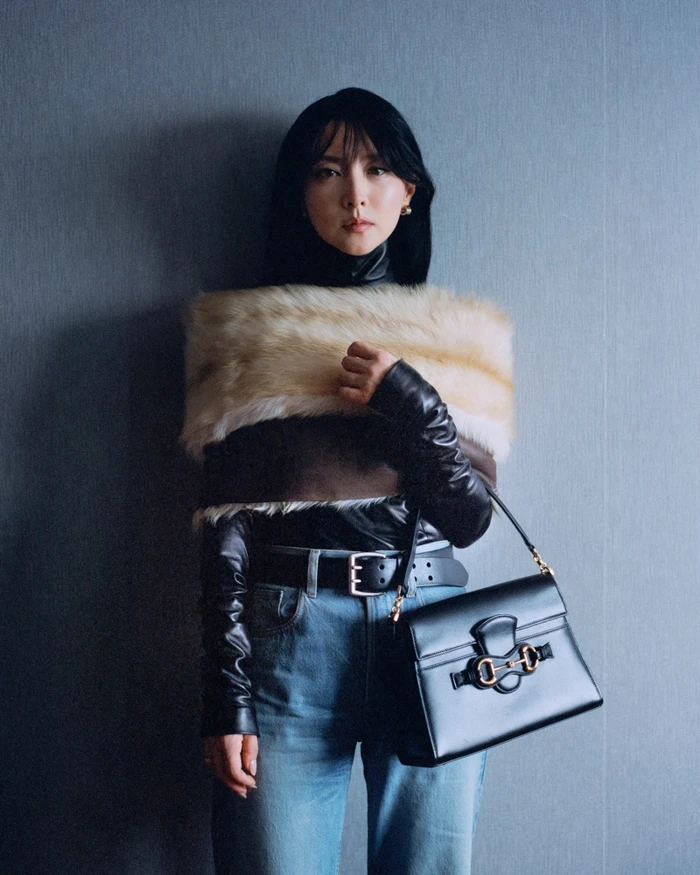 Lee Young Ae wearing a furry stole and black top with blue jeans and holding a handbag against a textured grey wall
