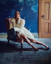Model Alex Consani seated in a pink velvet armchair wearing a cream fur coat against a textured blue wall for Gucci Primavera 2026.