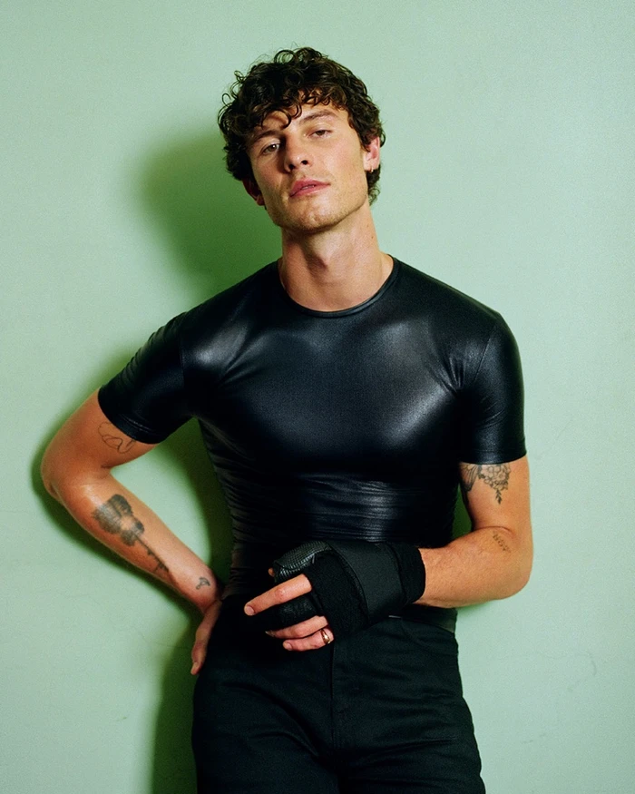 Portrait of Shawn Mendes wearing a black t-shirt and dark trousers against a pale green backdrop for Gucci Primavera 2026.