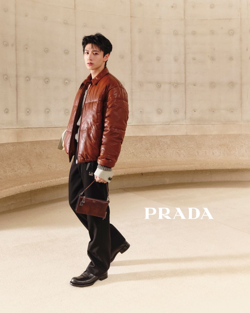 East Asian man in a brown quilted leather puffer jacket holding a small brown handbag against a curved concrete wall.