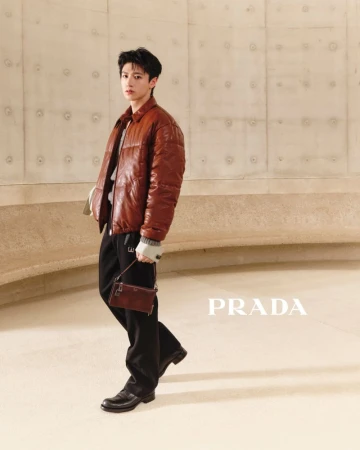 East Asian man in a brown quilted leather puffer jacket holding a small brown handbag against a curved concrete wall.