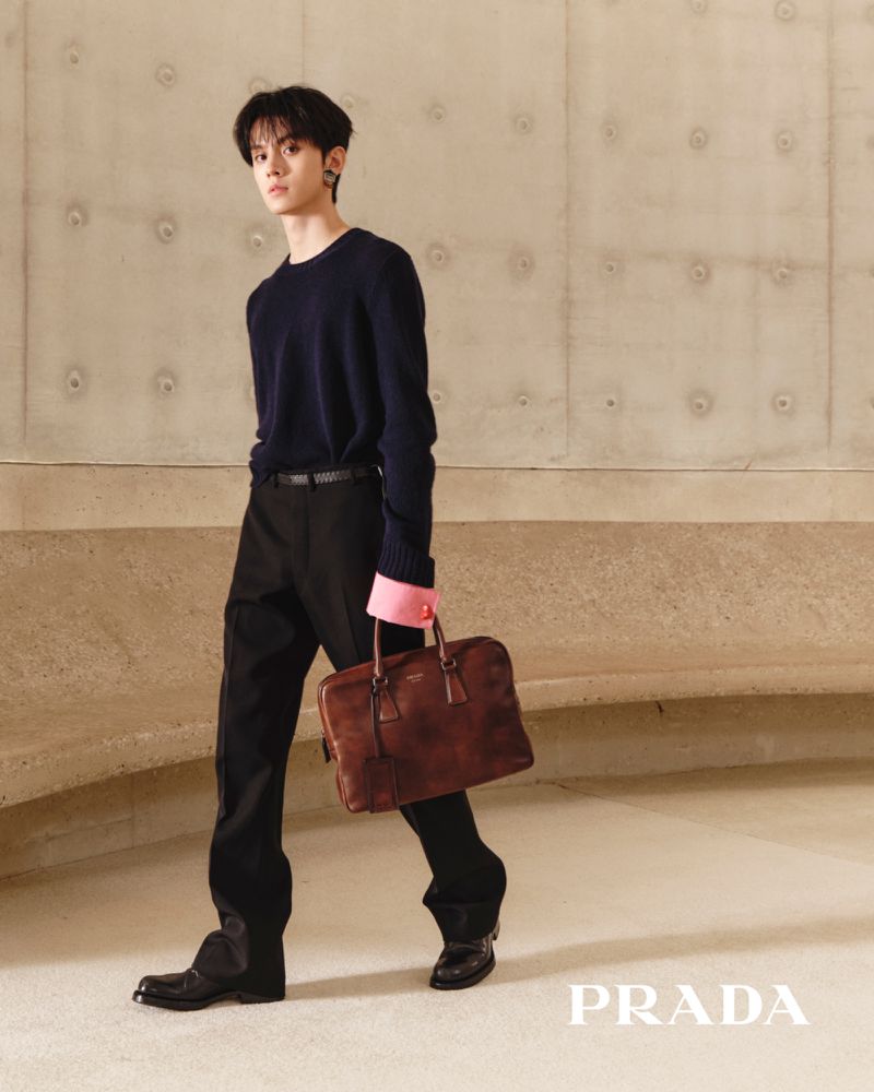 Win Metawin walks holding a brown leather briefcase, wearing a navy knit sweater and black trousers against a concrete wall.