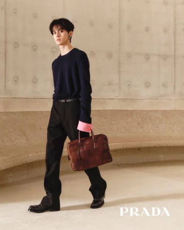 Win Metawin walks holding a brown leather briefcase, wearing a navy knit sweater and black trousers against a concrete wall.