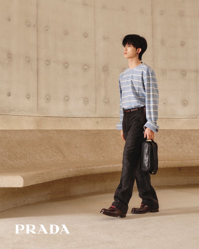 An East Asian male model walks holding a black leather bag, dressed in a blue striped shirt and dark trousers in a concrete space.