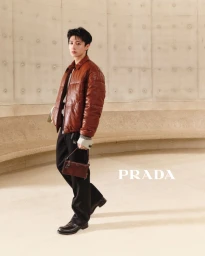 East Asian man wearing a brown leather puffer jacket and black trousers holding a small handbag against a curved architectural wall.