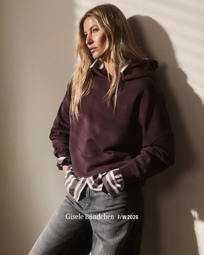 Gisele Bundchen wearing a purple hoodie layered over a striped shirt and jeans against a plain wall.