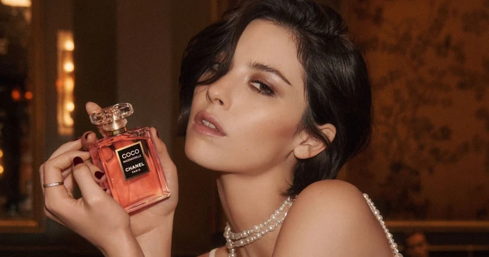 Gracie Abrams holding a Chanel Coco Mademoiselle perfume bottle wearing a pearl necklace in an indoor setting