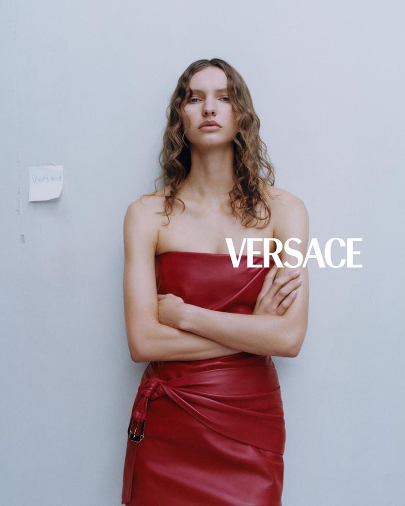 Fashion model in a red strapless top and wrap skirt, arms crossed, poses against a gray wall with Versace logo visible.