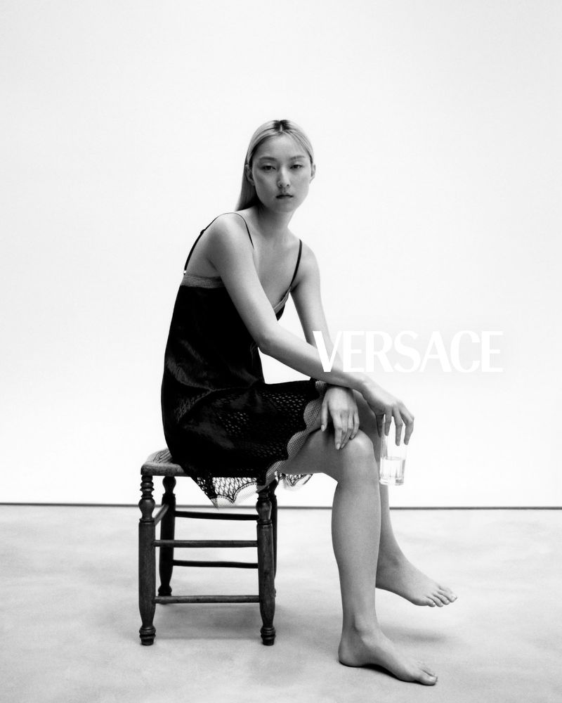 Elegant studio shot of a barefoot Asian woman in a black lace dress seated on a wooden stool, showcasing the VERSACE brand.