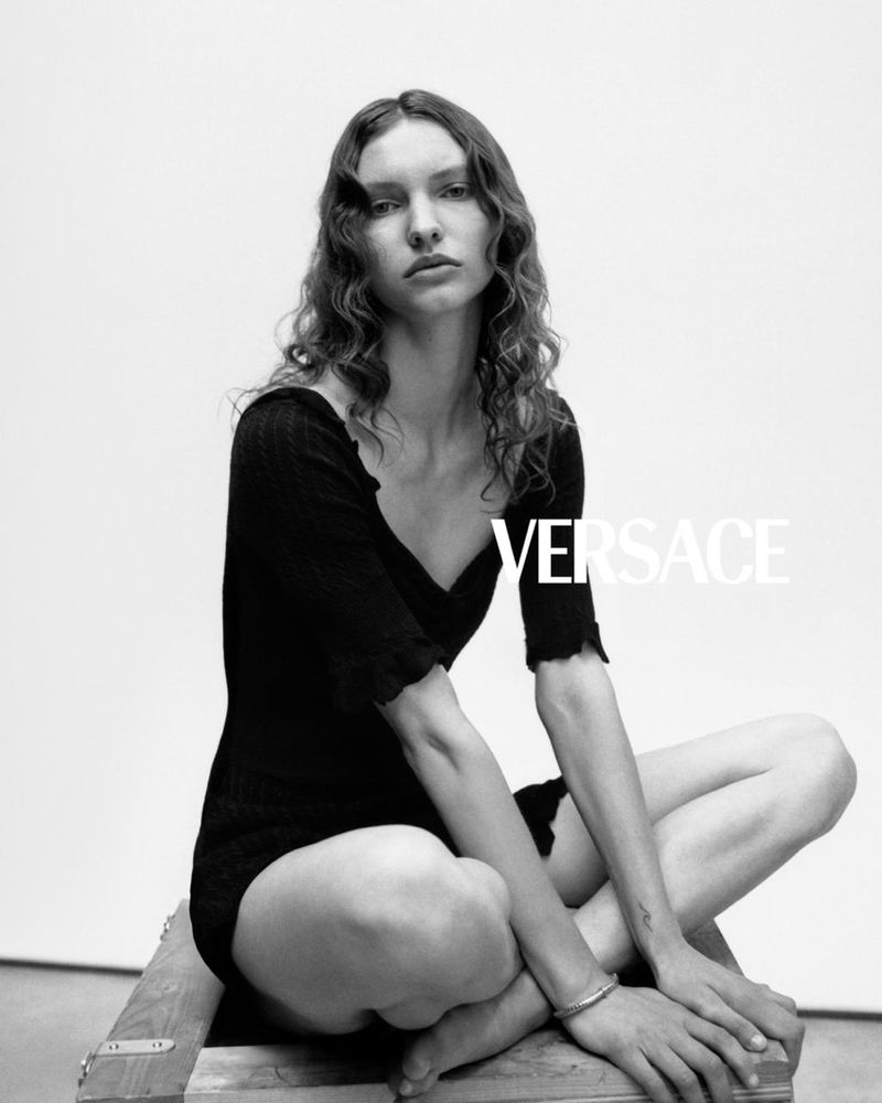 Monochrome portrait of a woman seated cross-legged on a wooden crate wearing a black short-sleeved dress against a white background.