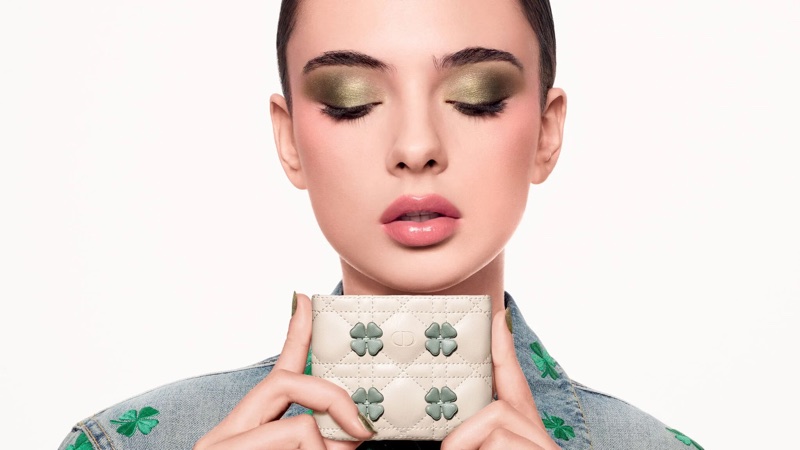 Close-up portrait of Deva Cassel holding a quilted white card holder with green clover details against a white background.