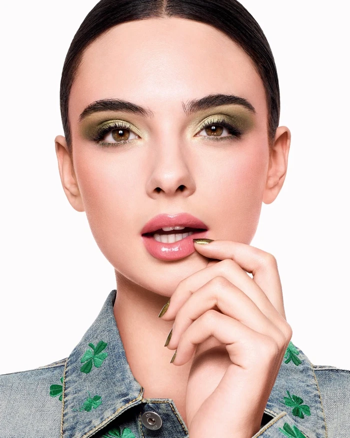 Close-up portrait of model Deva Cassel wearing olive green eyeshadow and a clover-patterned denim jacket against a white background.