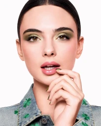Close-up portrait of model Deva Cassel wearing olive green eyeshadow and a clover-patterned denim jacket against a white background.