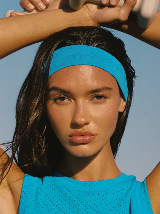 Woman wearing a bright blue headband and matching top against a clear blue sky, holding dumbbells behind her head.