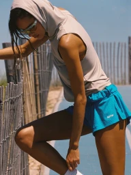 Woman wearing a beige hooded sleeveless top and bright blue athletic shorts leaning against a rustic wooden fence outdoors.