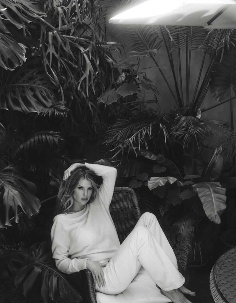 Charlotte McKinney reclining in a wicker chair wearing a white knit sweater and pants surrounded by tropical leaves in black and white.