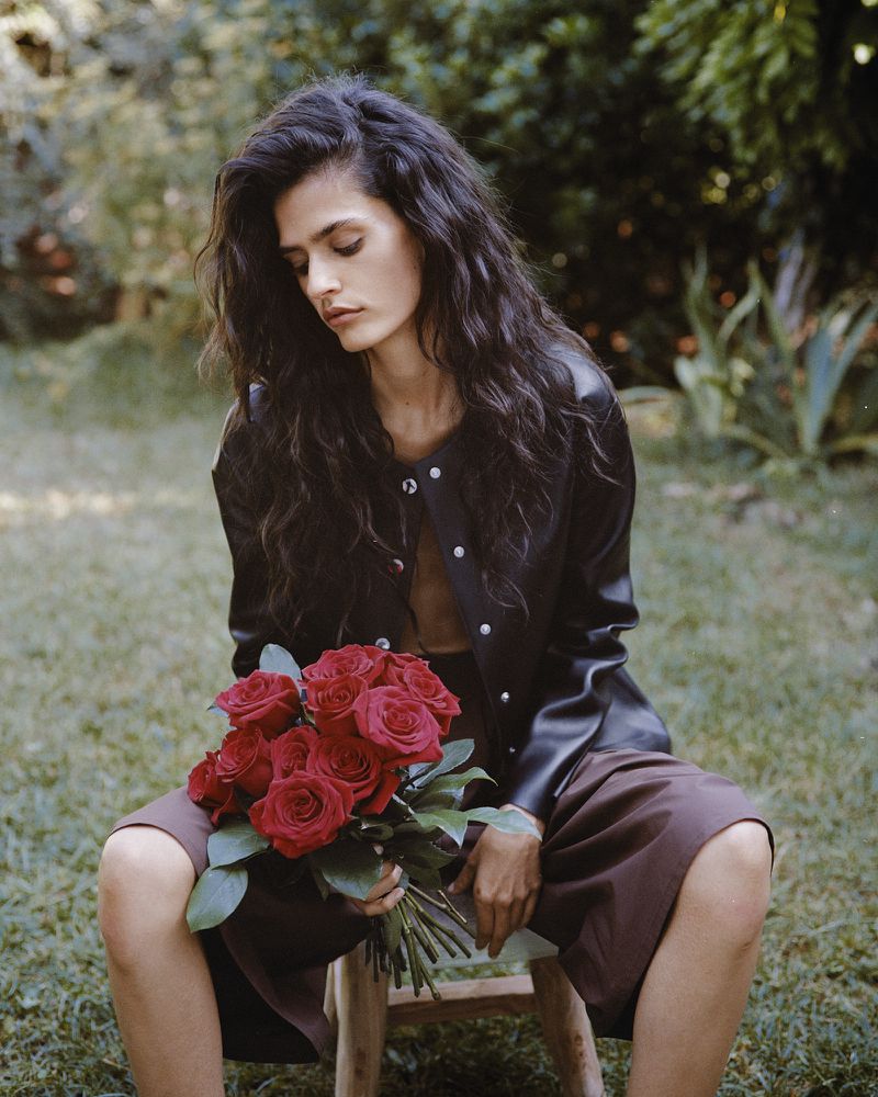 Avishag Amir wearing a black leather jacket and brown culottes while holding a bouquet of red roses outdoors.