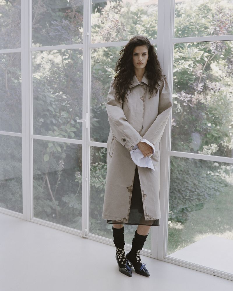 Model Avishag Amir standing in front of large floor-to-ceiling windows wearing a long beige trench coat and heels.