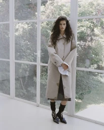 Model Avishag Amir standing in front of large floor-to-ceiling windows wearing a long beige trench coat and heels.
