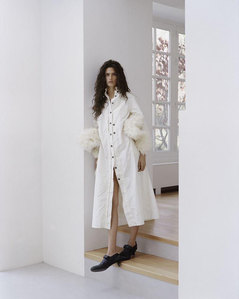 Model Avishag Amir in a long white button-down coat with feathered cuffs and black shoes standing on wooden steps indoors.