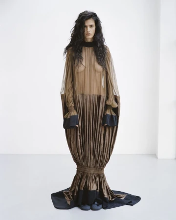 Avishag Amir posing in an avant-garde sheer brown pleated cocoon gown with black cuffs against a plain white studio background.