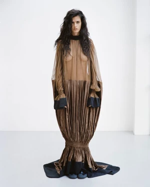 Avishag Amir posing in an avant-garde sheer brown pleated cocoon gown with black cuffs against a plain white studio background.