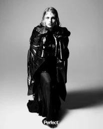 Model Karolina Spakowski standing in a glossy dark cape and pleated sheer skirt against a studio backdrop.