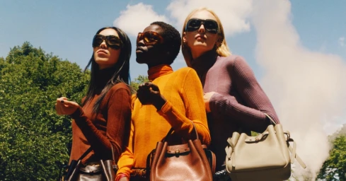 Three models wearing sunglasses and turtleneck sweaters posing outdoors against a clear sky with trees and smoke in the background.