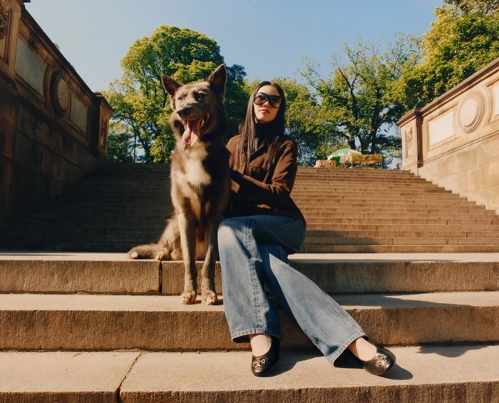 Model wearing dark sunglasses and brown jacket seated on stone steps beside a large dog under bright daylight