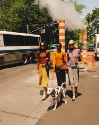 Three models walking a Dalmatian dog on a New York street next to a bus, wearing colorful knitwear and holding handbags.