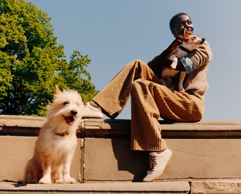 Model seated on stone wall wearing brown corduroy pants holding a small dog with another white terrier dog sitting below