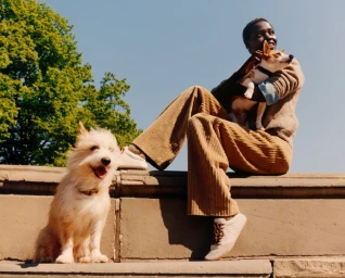 Model seated on stone wall wearing brown corduroy pants holding a small dog with another white terrier dog sitting below