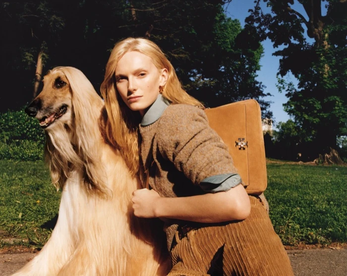 Model with long blonde hair kneeling outdoors beside an Afghan hound dog, wearing a brown sweater and carrying a tan suede handbag.
