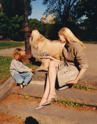 Model with long blonde hair holding a handbag and toy dinosaur on stone steps next to a child and a long-haired dog