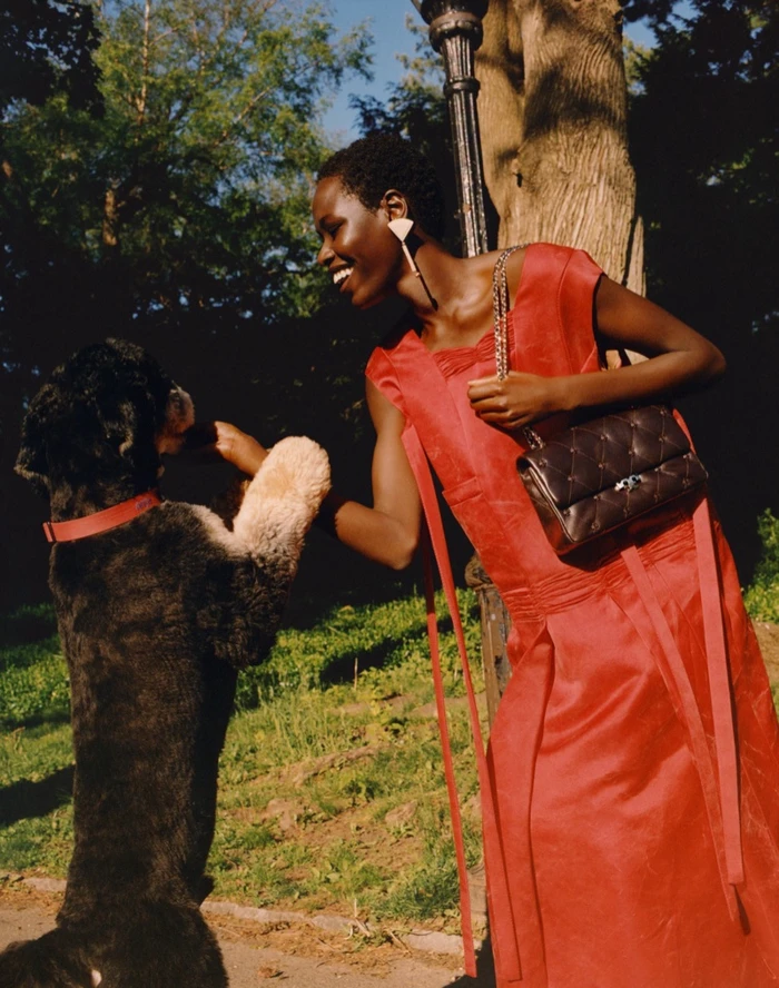 Model wearing a bright red sleeveless dress holding a quilted black handbag while interacting with a dog outdoors in a park.