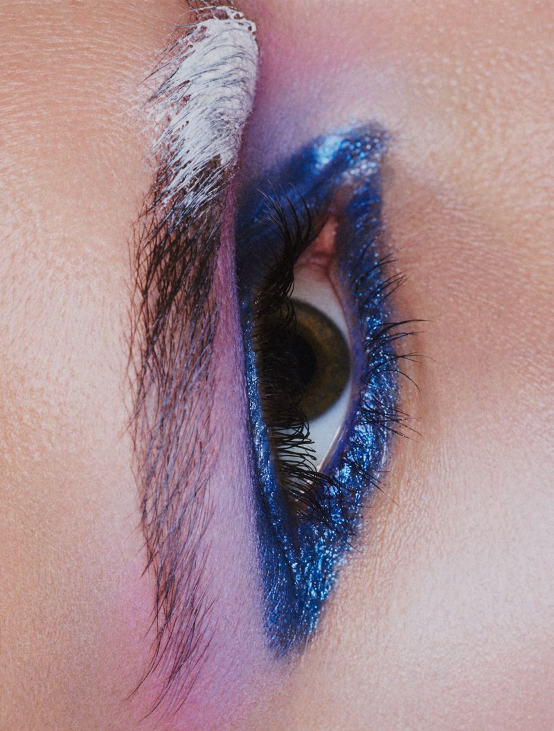 Extreme vertical close-up of a human eye with thick metallic blue eyeliner, a wash of purple eyeshadow, and an eyebrow partially painted white.