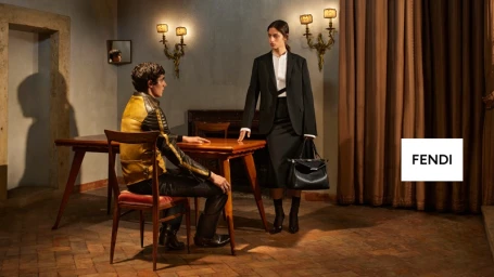 A man seated at a wooden table in a room and a woman standing beside him holding a black handbag for Fendi.