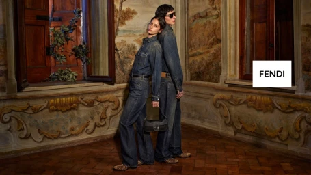 Two models wearing denim shirts and jeans lean back-to-back inside an ornate room with frescoed walls and wooden windows.